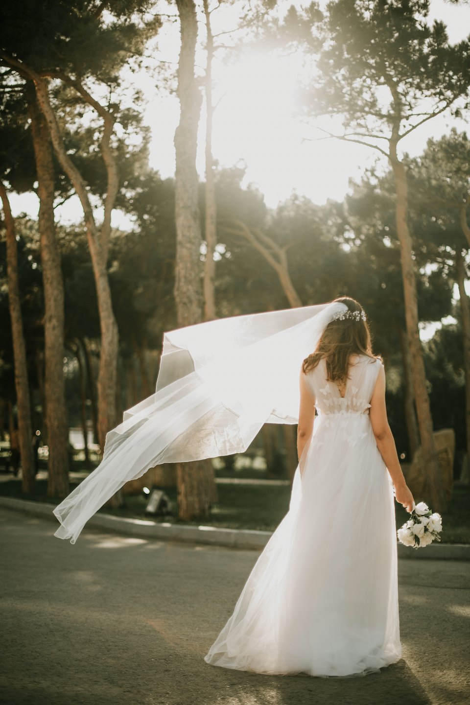 Sposa cammina lungo un sentiero fiancheggiato da pini con in mano dei fiori.